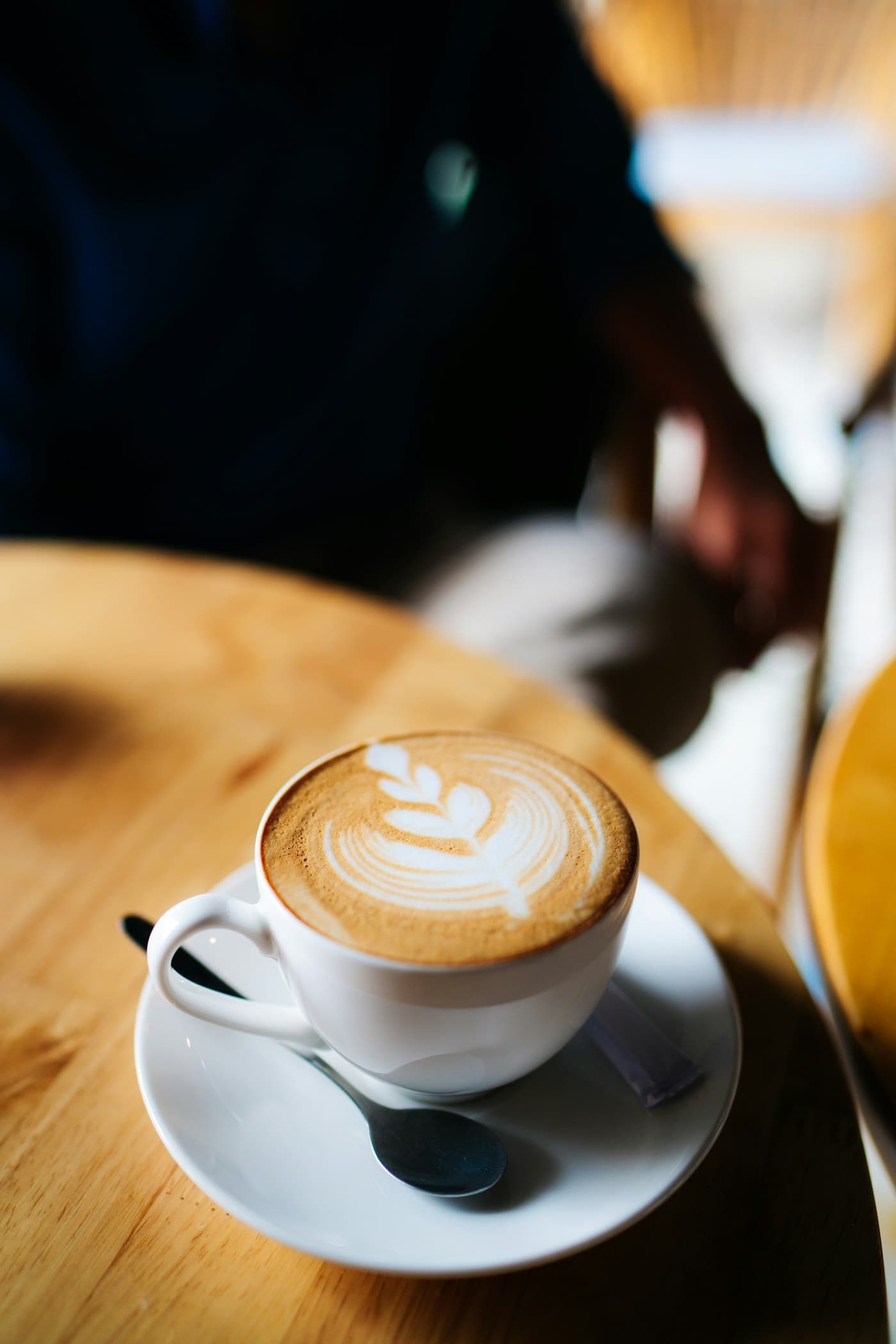 A cappuccino on a wooden table with a guest in soft focus behind